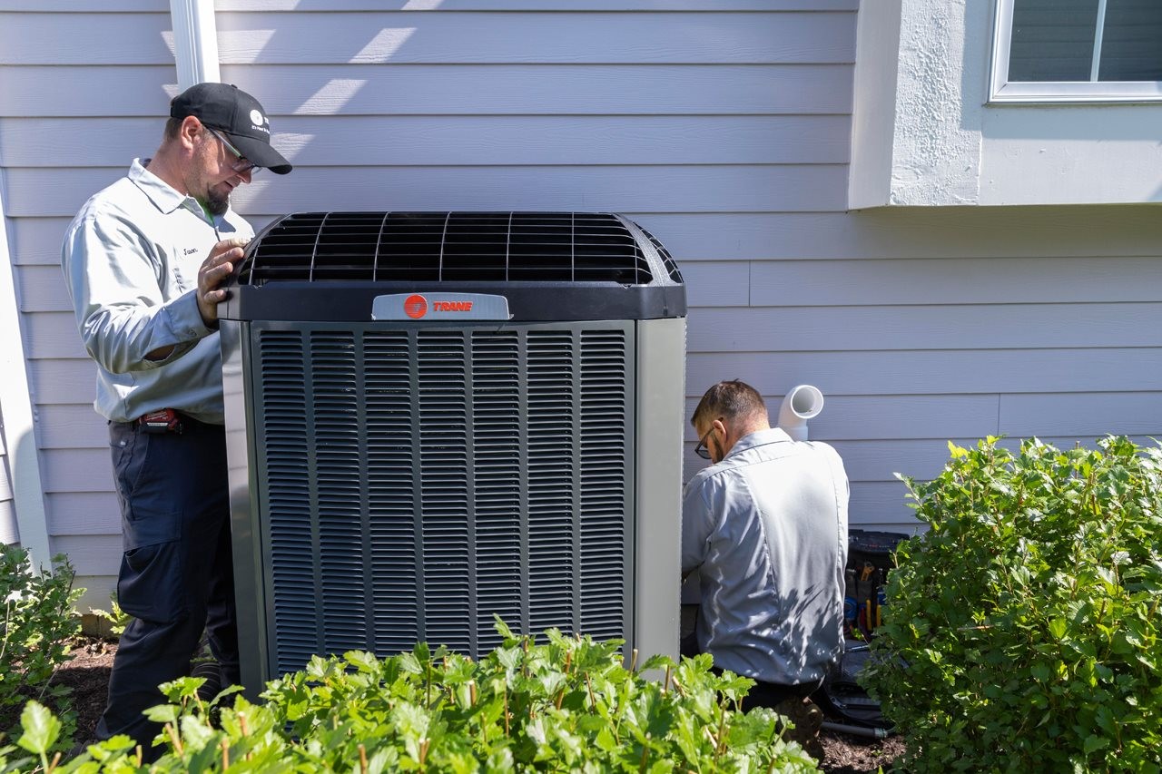 Two people installing a Trane HVAC system on th eoutside of a home.