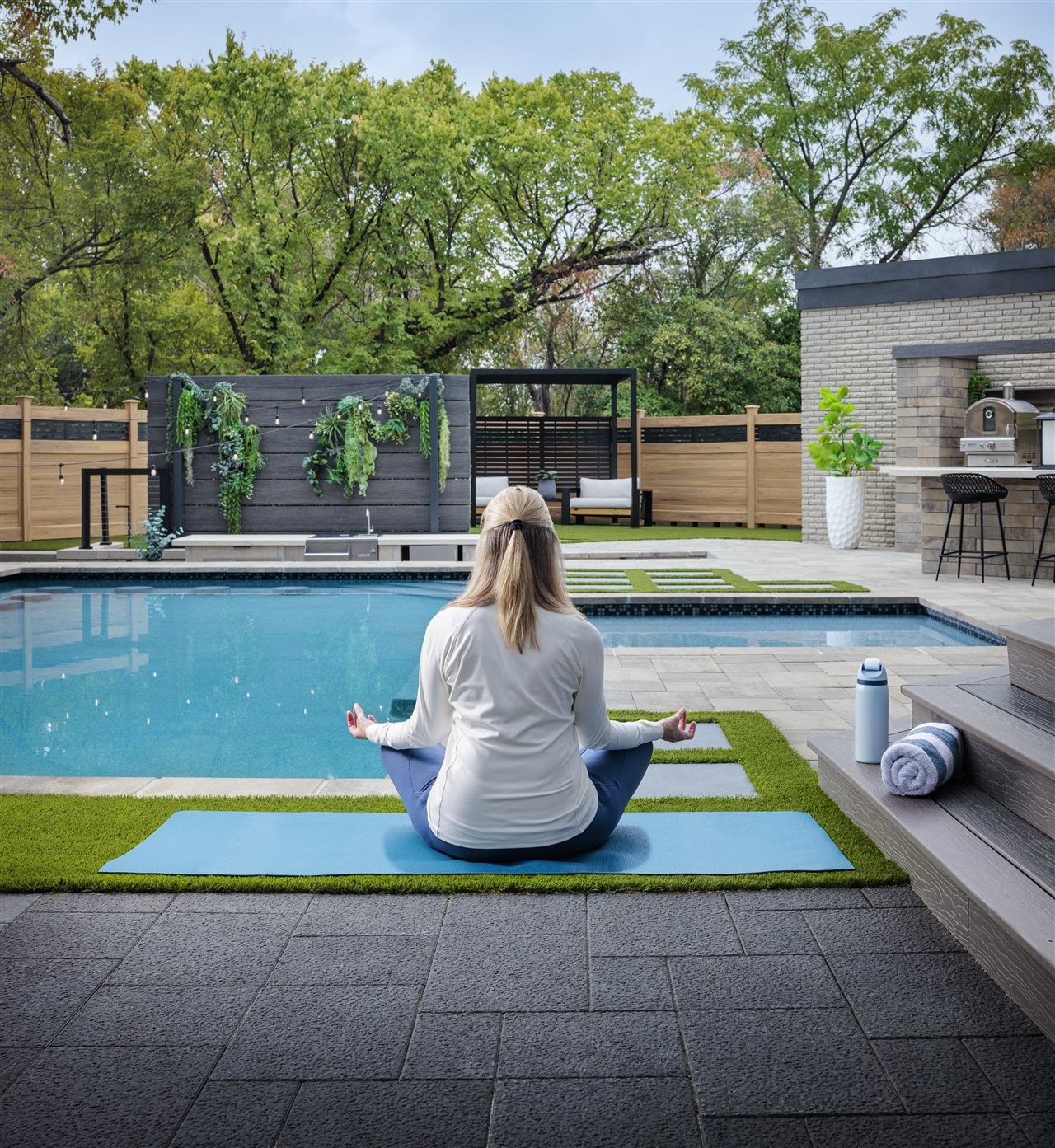 Woman praticing yoga on her back yard patio, in front of her pool, living wall and outdoor kitchen area.