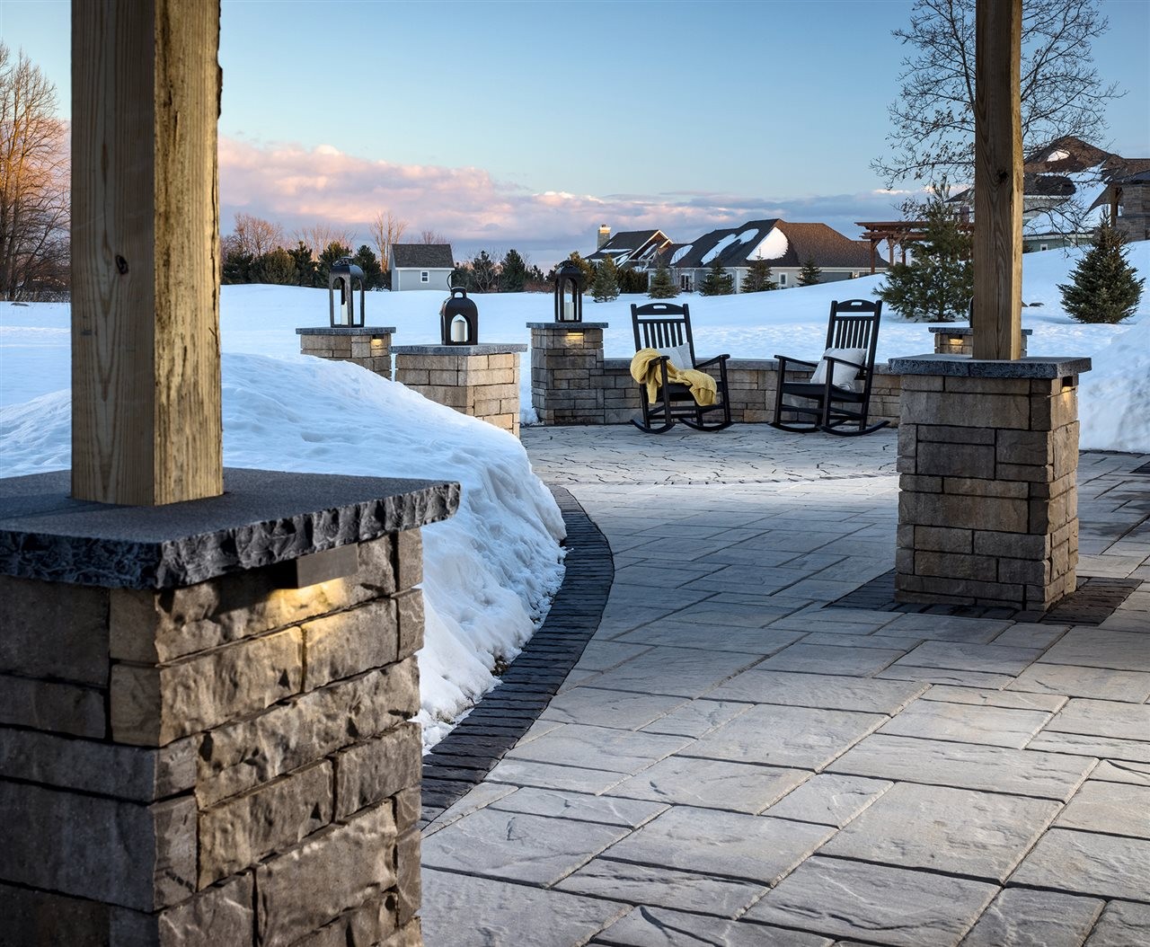 Paver patio path with lighted pergala pillars leaning to a sitting area with outdoor rocking chairs and cozy blankets.