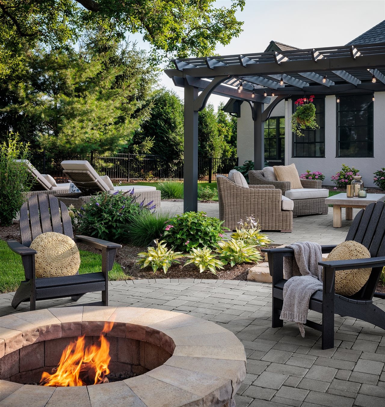 Lush landscaped backyard patio with pavers and comfy furniture under a pergala.